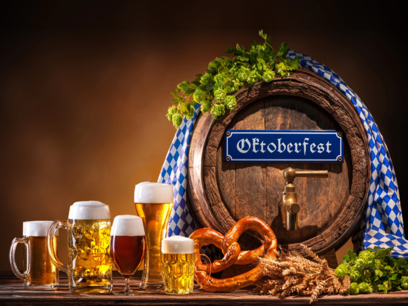 Das Bild zeigt ein Bierfass mit Schild Oktoberfest, daneben stehen verschiedene Biergläser mit unterschiedlichen hellen und dunklen Bieren gefüllt. Als Dekoration sind vorne eine Salzbretzel, ei n Strauß Getreideähren und grüne Hopfenranken dazugelegt.
