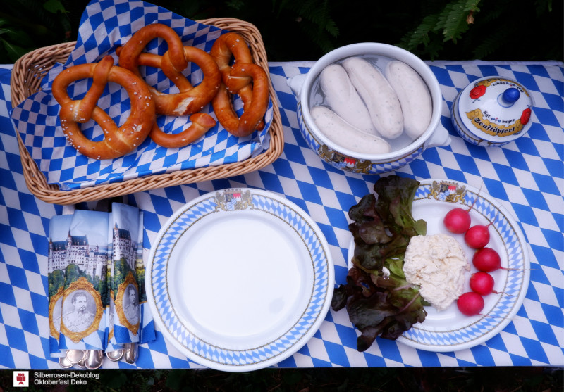 Der Tisch ist mit weiß-blauem Rautenmuster gedeckt. Darauf steht ein Brotkorb mit Laugenbrezeln, eine Schüssel mit Weißwürsten, ein Senftöpfchen, ein Teller mit Obatzda, Salatgarnitur und Radieschen sowie Geschirr und Besteck.