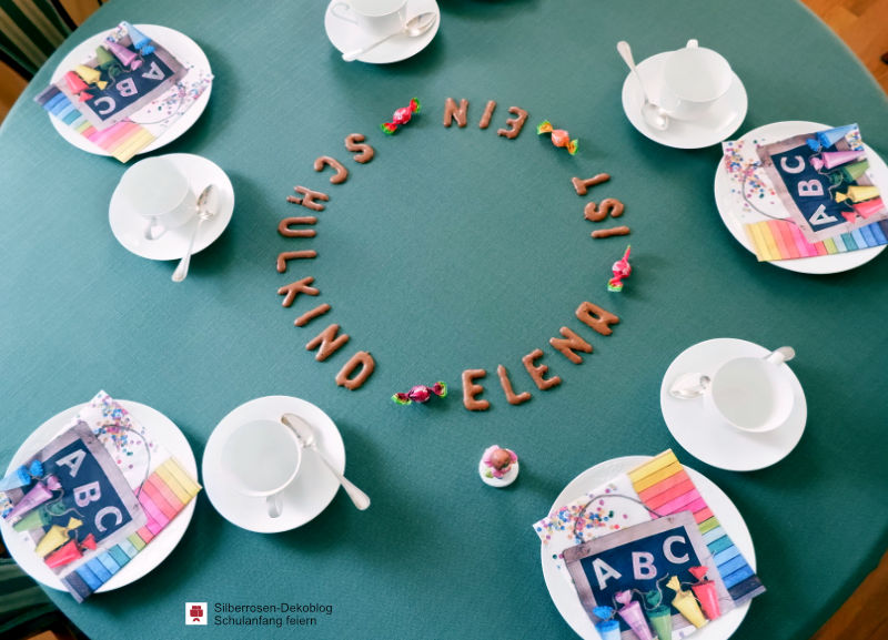Gedeckter Kaffeetisch für 5 Personen. In der Mitte ist mit Keksbuchstaben geschrieben: Elena ist ein Schuldkind. Auf den Tellern liegen bunte Einschulungsservietten mit einem Tafelmotiv, bunten Schultüten, Konfetti und einer Leiste bunter Rechtecke.