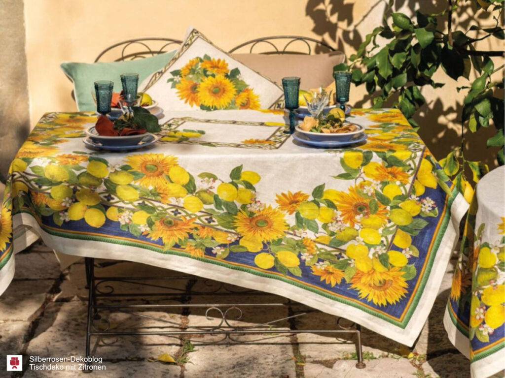 Ein Sitzplatz auf der Terrasse. Auf dem Tisch liegt eine Tischdecke, bedruckt mit einer breiten Bordüre aus Zitronen und Sonnenblumen vor blauem Hintergrund. Auf dem Tisch stehen Geschirr und farbige Gläser. Dahinter eine Bank mit Kissen.