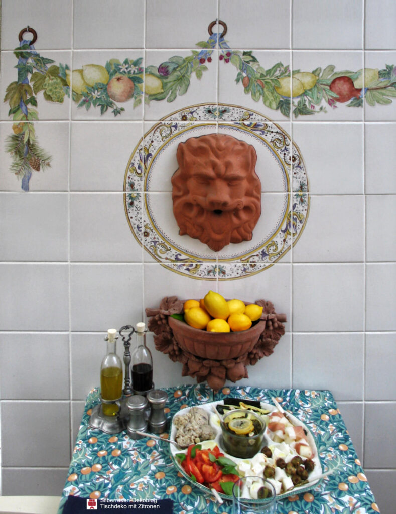 Bemalte Fliesenwand mit Fruchtgirlanden und Löwenmaske. Darunter eine Terracotta-Wandschale, gefüllt mit Zitronen. Davor steht ein gedeckter Tisch mit Essig &Öl, Salz & Pfeffer und Vorspeisenplatte.
