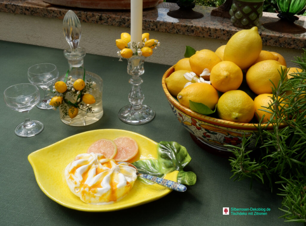 Auf einer grünen Tischdecke steht eine Keramikschale mit Zitronen, daneben ein Kerzenleuchter mit einem Zitronen-Kerzenkränzchen. Das gleiche Kerzenlkränzchen hängt als Deko um eine Karaffe mit Limoncello. Davor steht ein Teller in Form einer Zitrone mit Dessert.