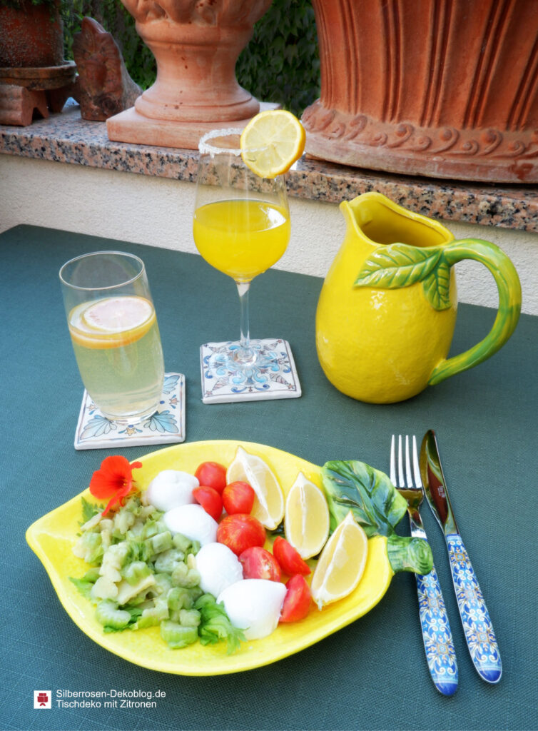 Auf dem Tisch steht ein Limonadenkrug in Form einer Zitrone, zwei Gläser mit Getränken und Zitronenscheiben und ein Teller in Zitronenform mit Salat, Mozzarellabällchen und Kirschtomaten sowie Zitronenspalten.