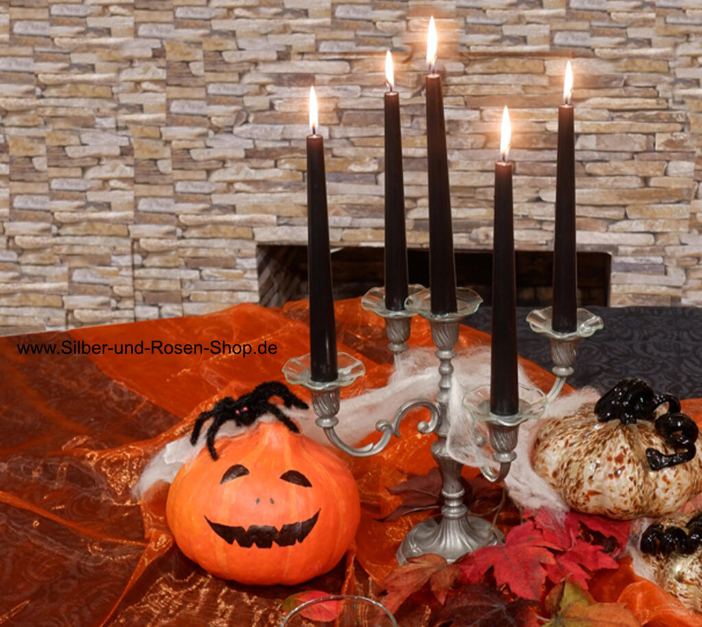 gedeckter Halloween-Tisch mit einem Kürbis, dem ein Gesicht aufgemalt wurde, dazu ein Kerzenleuchter mit schwarzen Kerzen und orangefarbene Tischdecke mit Herbstlaub.
