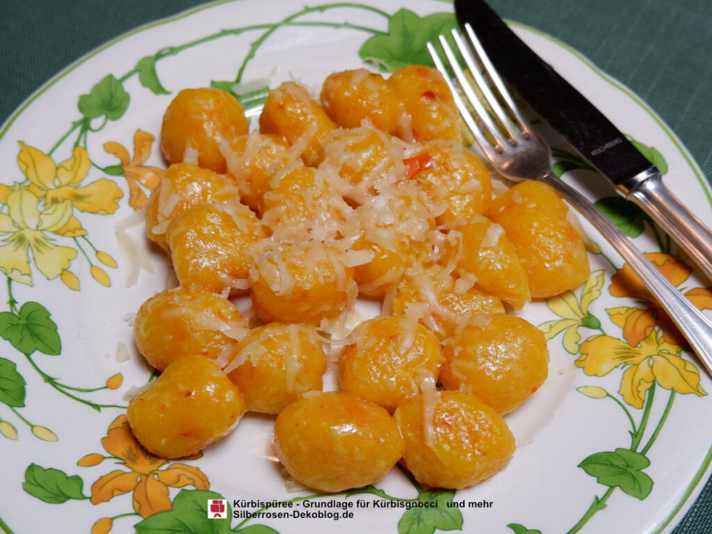 Goldgelbe Kürbisgnocci aus dem Rezept auf einem Teller mit Parmesam bestreut. Der Teller hat Ranken von grünen Blättern und gelben Blüten. Daneben liegt Besteck.