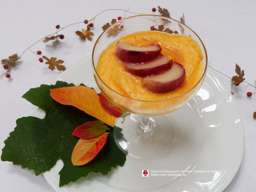In einem Dessertglas ist Kürbismousse angerichtet mit karamellisierten Pflaumen. Auf dem Teller darunter liegen Herbstblätter. Eine Metallblättergirlande liegt außen um den Teller.