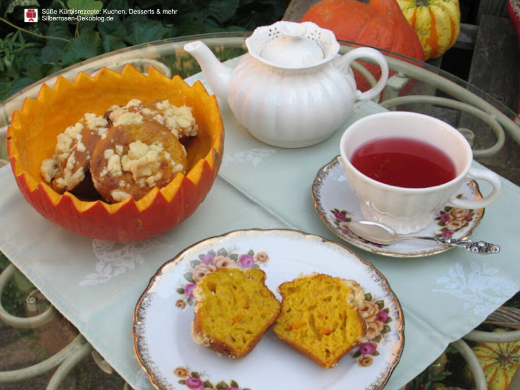 Auf einem kleinen Gartentisch sind Kürbismuffins in einer ausgehöhlten Kürbisschale serviert. Auf dem Teller daneben ist ein Muffin aufgeschnitten. Eine gefüllte Teetasse und ein Teekännchen stehen dabei.
