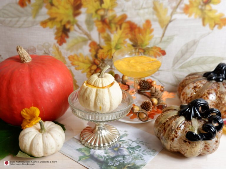 Süße Kürbisrezepte angerichtet. Vor bunten Herbstblättern liegen weiße und orange Kürbisse, dazwischen steht ein Kelch mit einem Kürbisdessert. In einem kleinen weißen Kürbis ist ebenfalls Kürbisdessert.