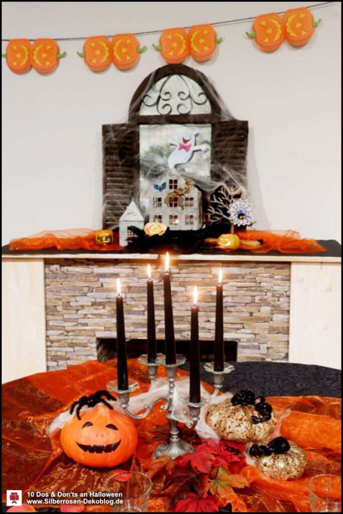 Dekorierte Halloween Szene. Im Hintergrund ein Kamin mit aufgebauter Deko: Spiegel mit Spinneweben, Halloween-Sticker. Im Vordergrund ein gedeckter Tisch: oranges Organzamit fünfarmigem Kerzenleuchter mit schwarzen Kerzen. Daneben Kürbis mit Spinne und Glaskürbisse.