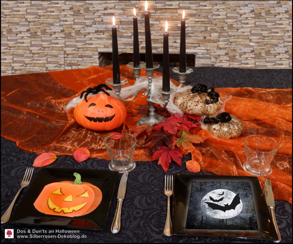 Gedeckter Halloweentisch mit orangem Organza und Kerzenleuchter mit schwarzen Kerzen. Im Vordergrund zwei schwarze Teller mit unterschiedlichen Servietten.