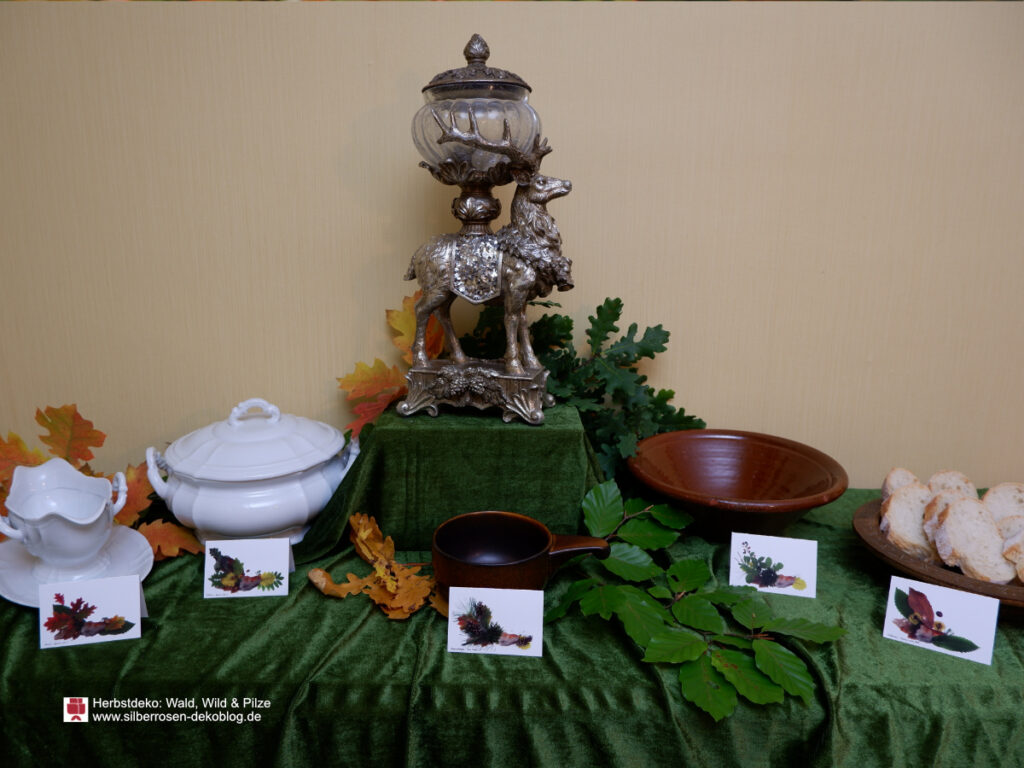 Probedecken des Herbstbuffets. Grüne Tischdecke. Auf einem Podest in der Mitte eine Hirschfigur, darum Herbstlaub. Schüsseln, deren Inhalt auf Platzkarten davor steht.