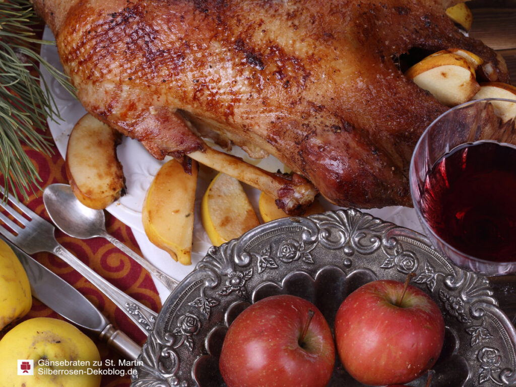 Das Bild zeigt eine gebratene Gans auf einer Platte mit Apfelstücken. Daneben eine Schale mit roten Äpfeln, ein Glas Rotwein, Besteck und Quitten.