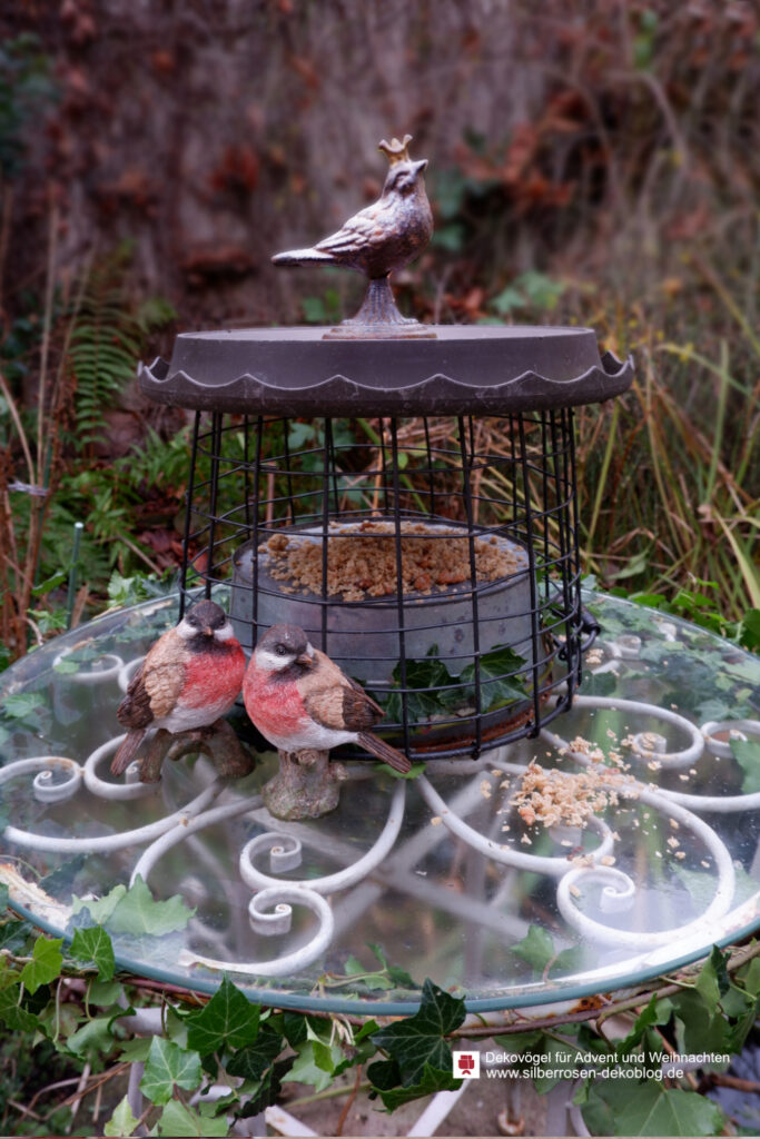 Geschützter Futterplatz für Vögel in einem Gitterkorb auf einem Gartentisch. Als Deko stehen zwei kleine Polyresin-Vögel daneben.