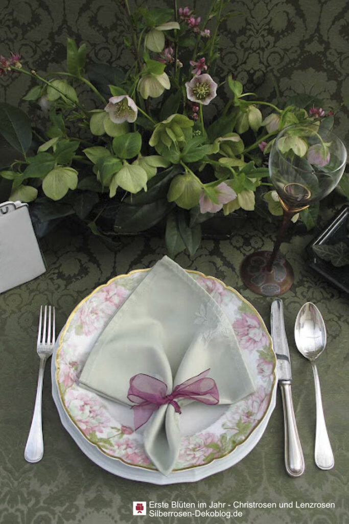Blumengesteck mit rosa und grünen Lenzrosen sowie blühenden rosa Pfirsichzweigen. Davor ein Gedeck mit rosa-grün gemustertem Teller.