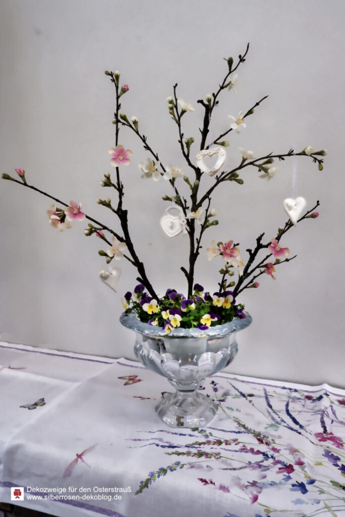 In einer Glasschale mit Stiefmütterchen stecken künstliche Osterzweige mit rosa und weißen Blüten.