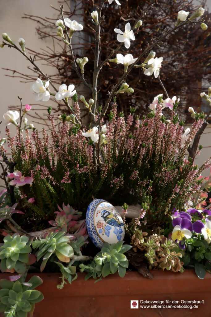 Osterstrauß mit künstlichen Blütenzweigen. Künstliche Zierquittenzweige mit weißen und pinken Blüten stecken in einem bepflanzten Balkonkasten mit Heidekraut und Stiefmütterchen. Darin liegt ein Osterei.