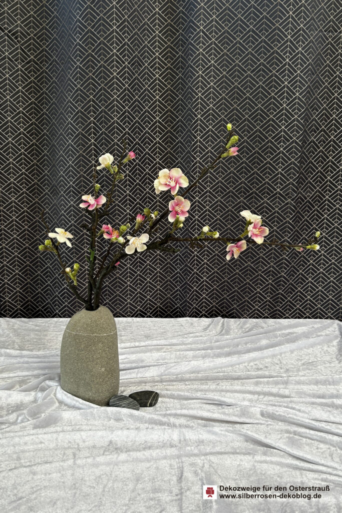 Ein künstlicher Zierquittenzweig mit rosa-weißen Blüten steckt in einer Kieselsteinvase. Das Tuch darunter ist wie fließendes Wasser in Falten gelegt und staut sich ein wenig an der Vase. Zwei kleine Kieselsteine liegen daneben.