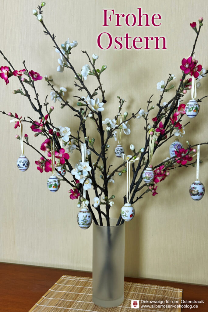 Mehrere künstliche Zierquittenzweige mit weißen und pinken Blüten stehen als Osterstrauß in einer glatten Glasvase. An den Zweigen hängen kleine Hutschenreuther Porzellaneier.