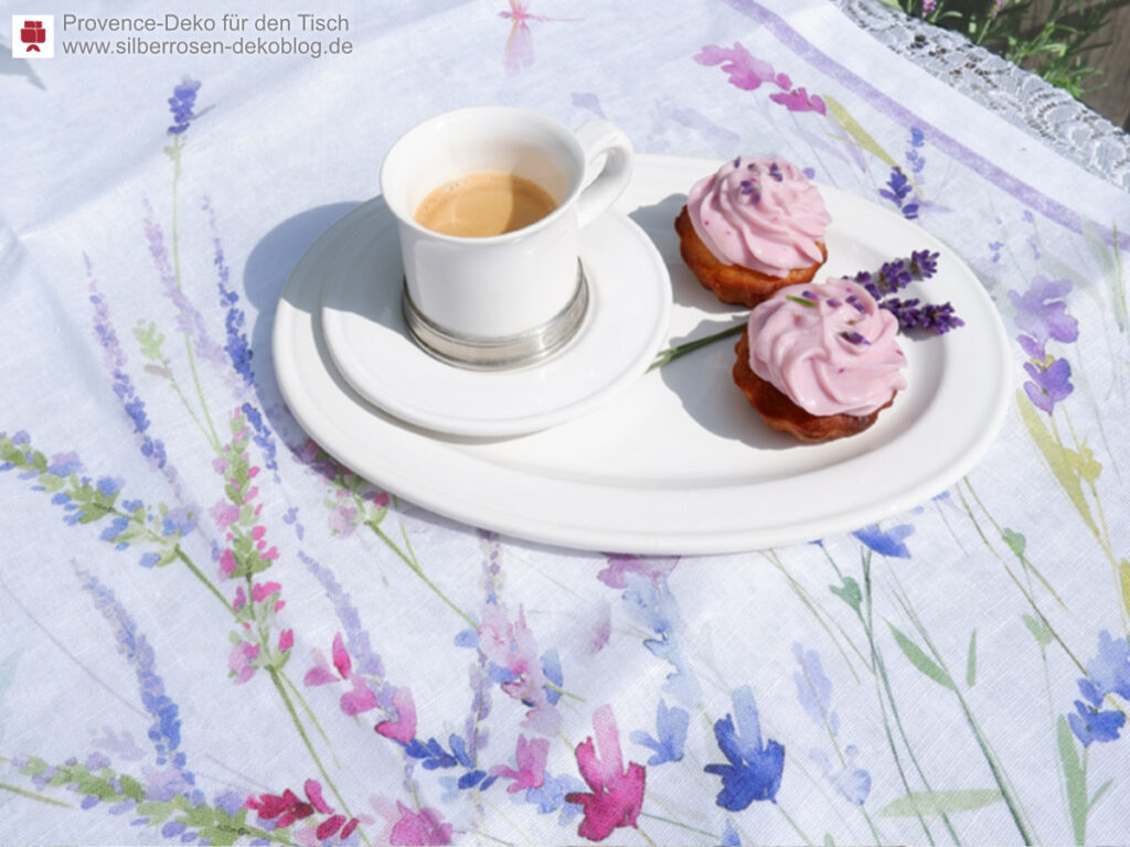 Auf einer Tischdecke Spigo mit Lavendelblüten steht eine ovale weiße Platte mit einem Espresso und zwei lila Cupcakes. 